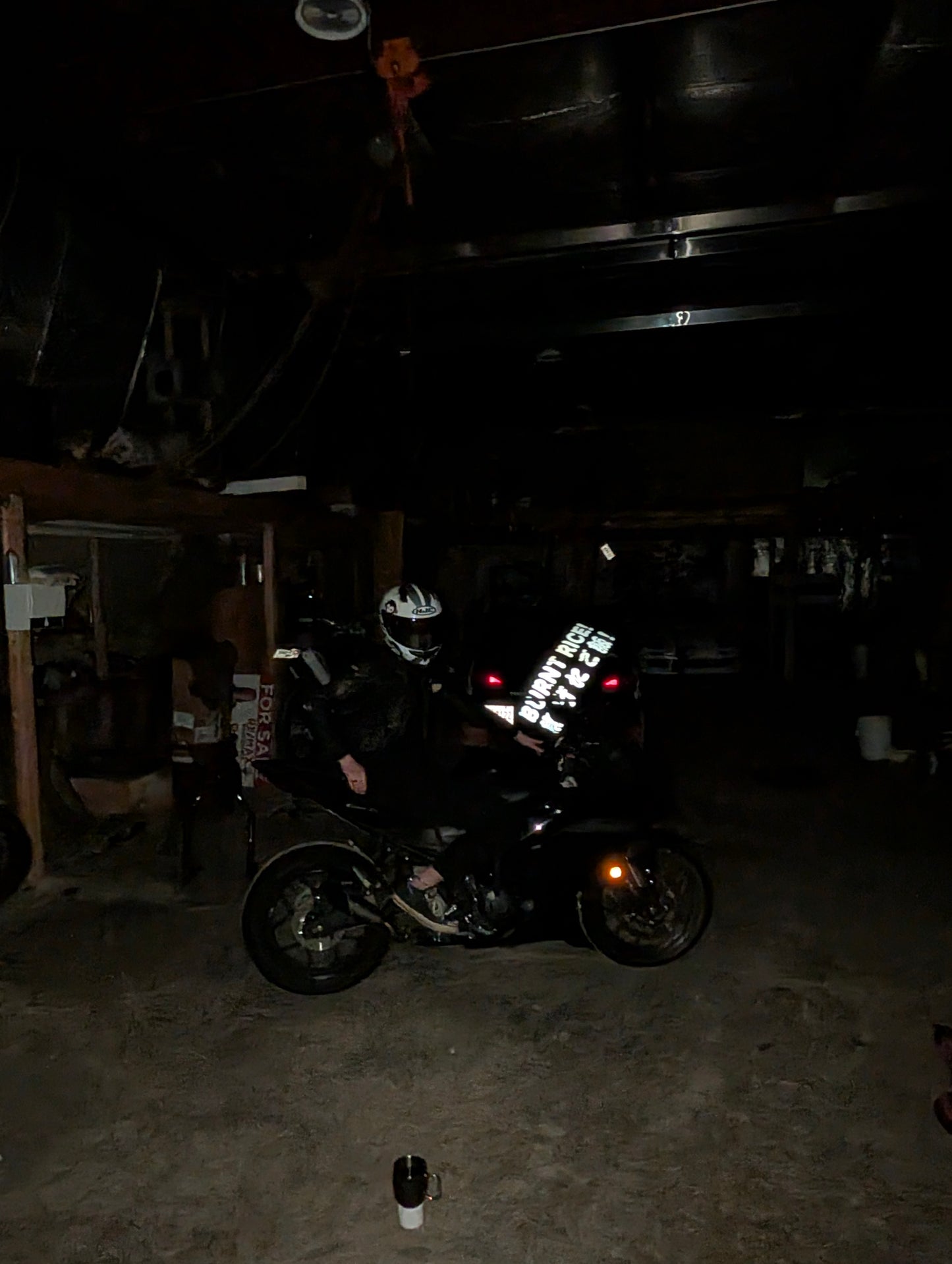 A black motorcycle with a rider wearing a white helmet, parked inside a dark garage. The motorcycle has a prominent white graphic decal on the side and a matching white graphic on the windshield.