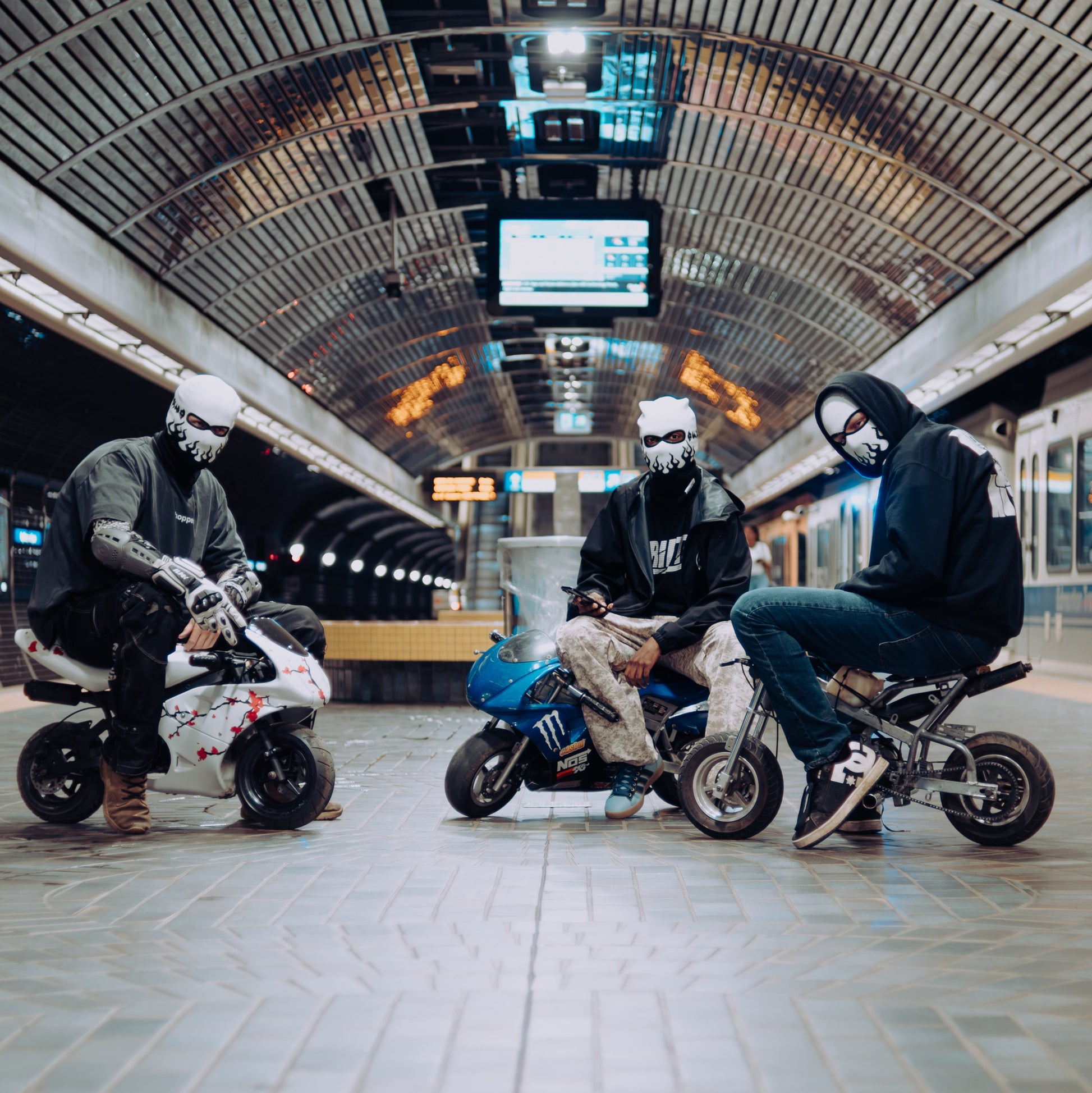 Three people wearing face masks sit on mini motorbikes in a modern, arched subway station with tiled floors and overhead lights.