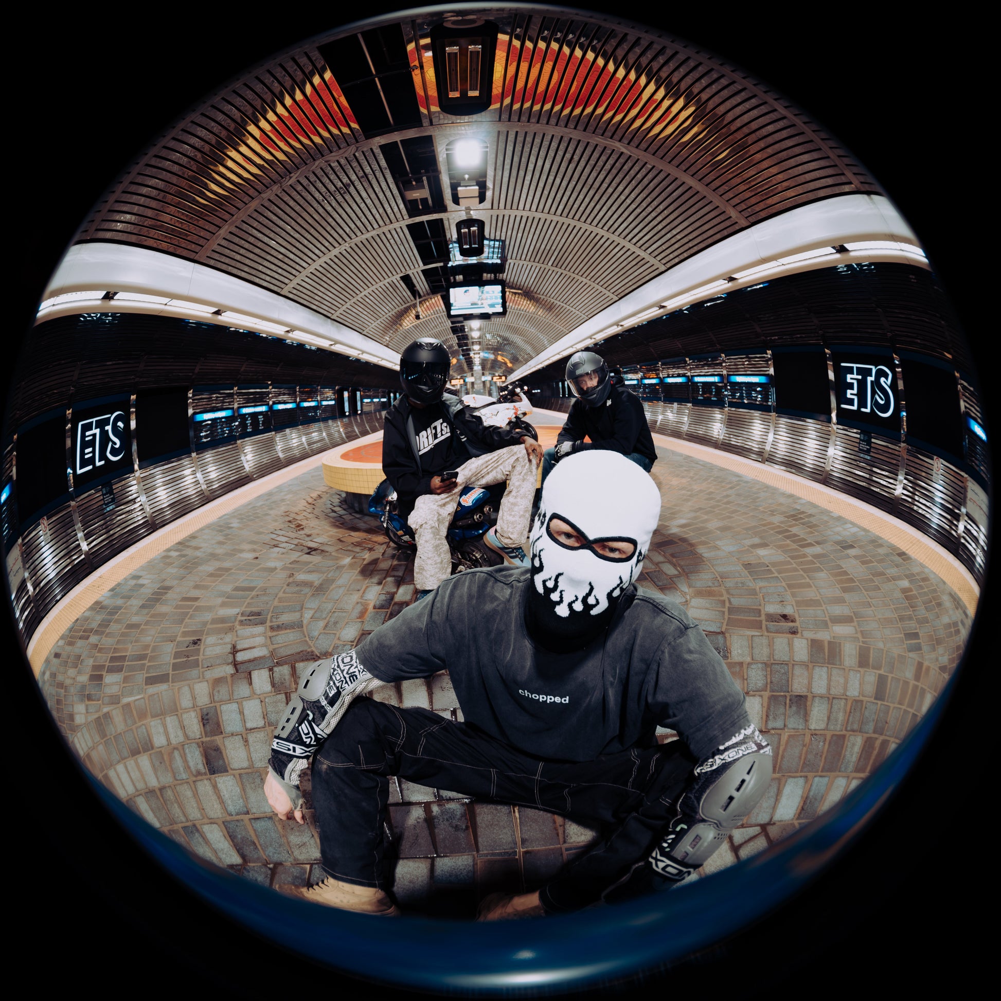 A person wearing a white and black face mask and a dark gray hoodie sits in a subway station, captured through a fisheye lens. Other people are visible in the background of the station, and the image is framed by a black circular border.