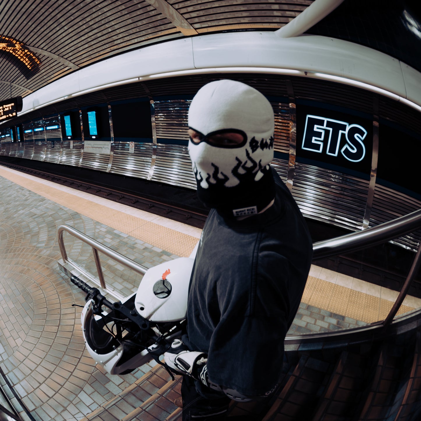 A person wearing a white balaclava mask with black graphic print, a black hoodie, and white gloves stands on a subway platform holding a white helmet. The photo uses a dramatic fisheye lens effect, showing a curved station interior with a visible 'ETS' sign in the background.