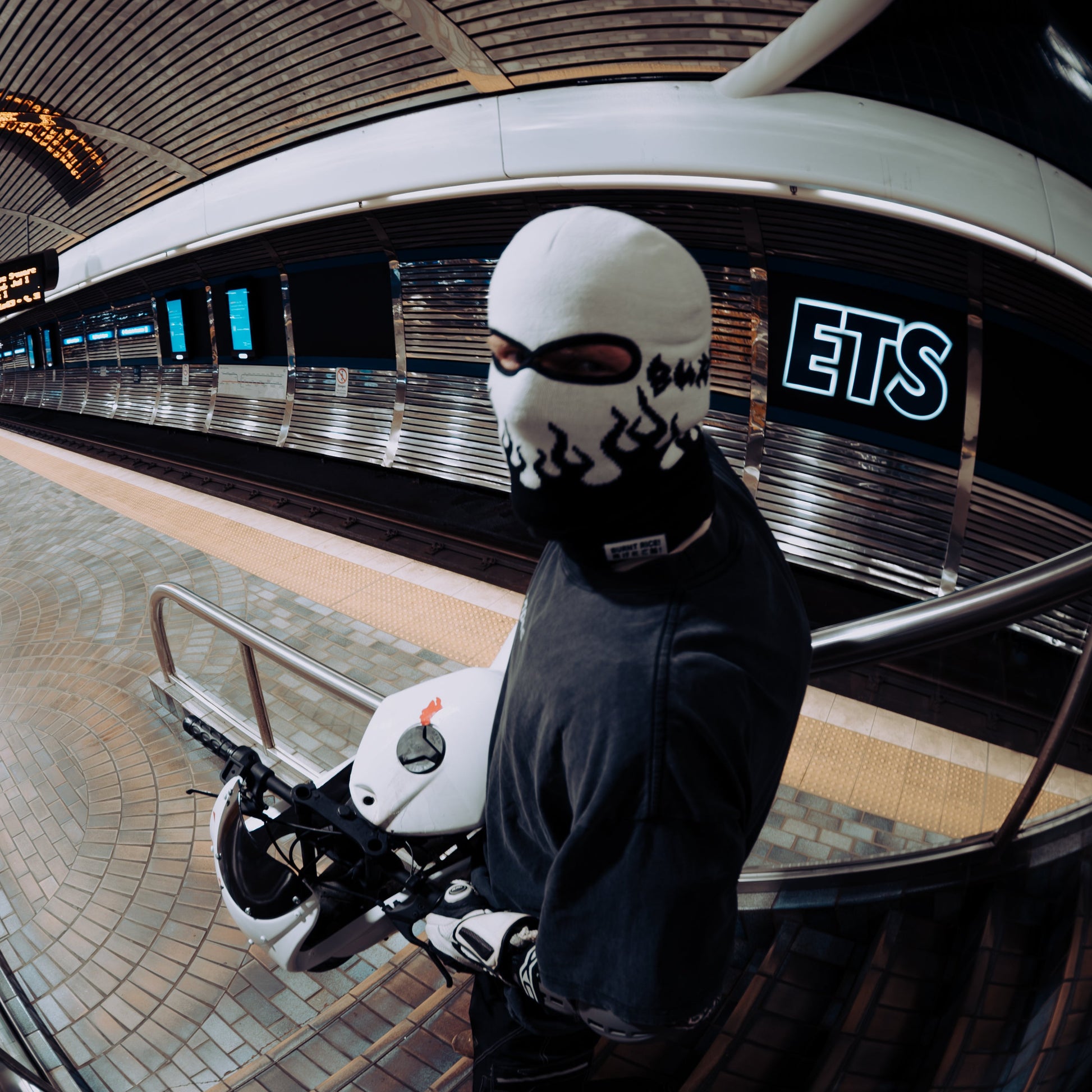 A person wearing a white balaclava mask with black graphic print, a black hoodie, and white gloves stands on a subway platform holding a white helmet. The photo uses a dramatic fisheye lens effect, showing a curved station interior with a visible 'ETS' sign in the background.
